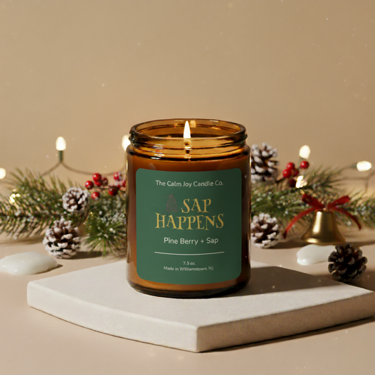 Candle labeled 'Sap Happens' from The Calm Joy Candle Co. on a decorative surface with Christmas decorations.