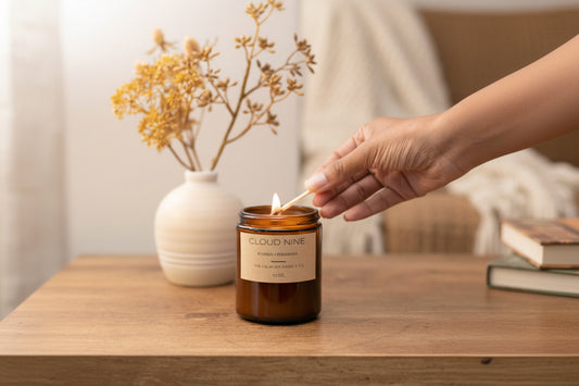A person's hand lighting a Pumpkin Persimmon Candle with a match, which is placed on a wooden surface, with a vase of dried flowers in the background.