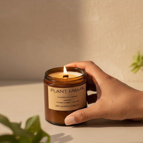 Hand holding a 'Plant Mama' candle with plants in the background