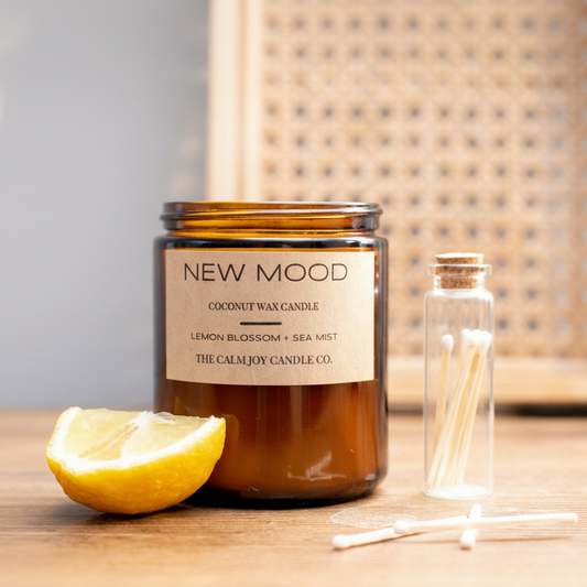 A brown jar of 'NEW MOOD' coconut wax candle with lemon blossom and sea salt mist fragrance, accompanied by a clear glass holder with white wax tabs and a lemon slice in the foreground.